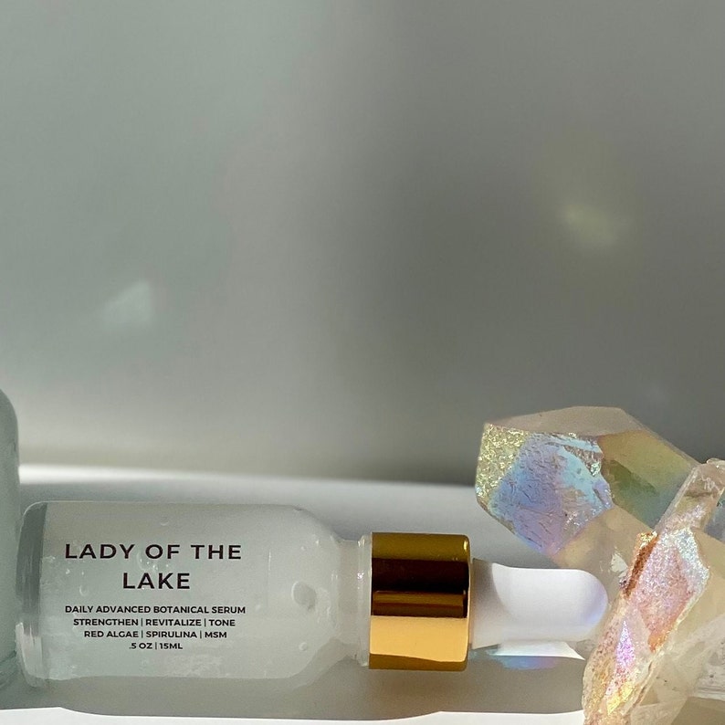 May include: A white bottle of "Lady of the Lake" daily advanced botanical serum. The bottle has a gold-colored cap and a white dropper. The label lists ingredients like red algae and spirulina. The background includes crystals.
