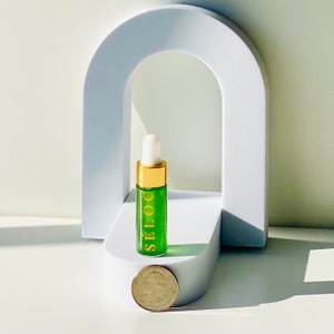 K&ouml;nnte beinhalten: Eine kleine gr&uuml;ne Glasflasche mit goldener Kappe und Tropfer. Die Flasche ist mit "SELOC" in goldenen Buchstaben beschriftet. Die Flasche steht auf einer wei&szlig;en Plattform mit einer US-amerikanischen 25-Cent-M&uuml;nze davor.