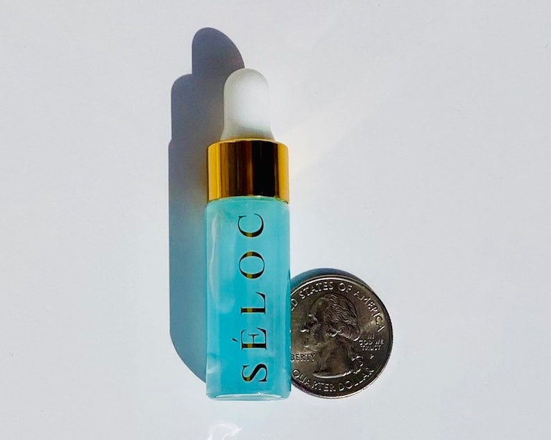Pode incluir: Um pequeno frasco de vidro transparente com uma tampa dourada e um l&iacute;quido azul dentro. O frasco &eacute; rotulado com "S&Eacute;LOC" em letras douradas. O frasco est&aacute; ao lado de uma moeda de 25 centavos de d&oacute;lar americano.