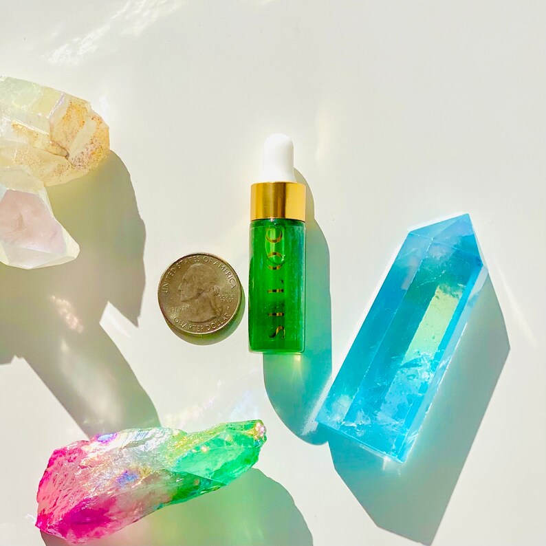 K&ouml;nnte beinhalten: Eine kleine gr&uuml;ne Glasflasche mit goldenem Deckel und einem wei&szlig;en Tropfer. Die Flasche ist mit "SERUM" beschriftet. Die Flasche steht auf einer wei&szlig;en Oberfl&auml;che mit einem blauen Kristall, einem rosa und gr&uuml;nen Kristall und einem wei&szlig;en Kristall.