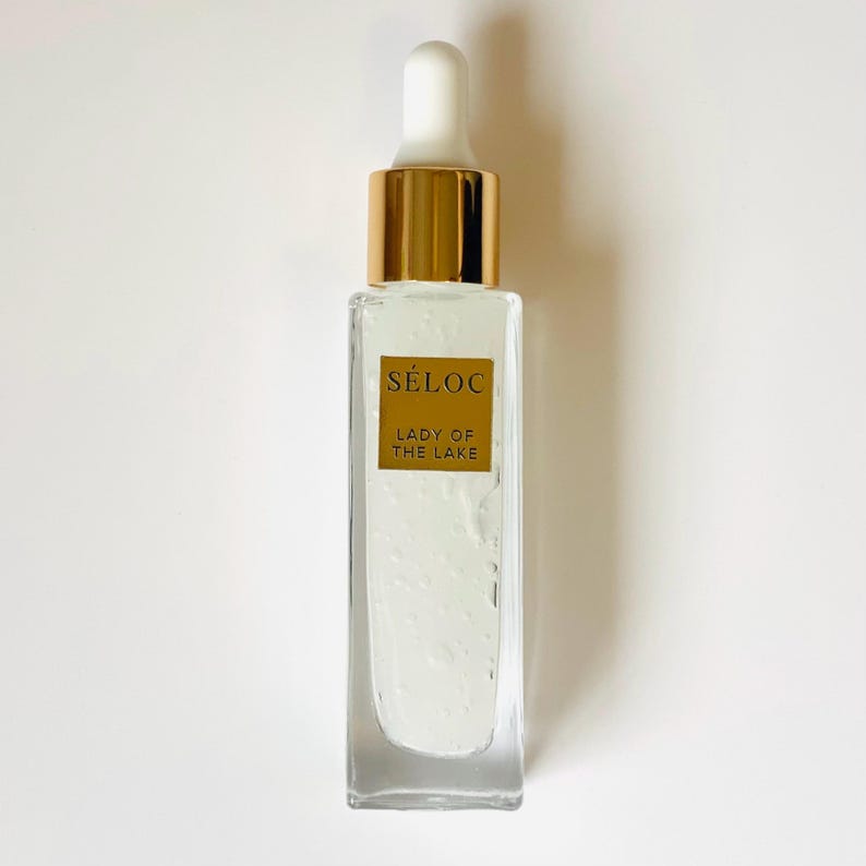 May include: A clear glass bottle with a white dropper and gold-colored cap. The bottle contains a clear liquid and has a gold label with the text "S&Eacute;LOC LADY OF THE LAKE". The bottle is rectangular and sits on a white surface.