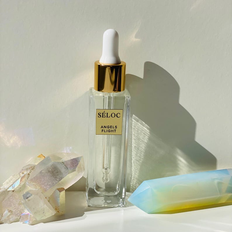 K&ouml;nnte beinhalten: Eine klare Glasflasche mit goldenem Verschluss und wei&szlig;er Pipette, beschriftet mit "S&Eacute;LOC ANGELS FLIGHT". Die rechteckige Flasche enth&auml;lt eine klare Fl&uuml;ssigkeit. Neben der Flasche befinden sich zwei Kristalle, einer klar und einer blau und gelb.