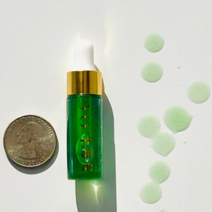 K&ouml;nnte beinhalten: Eine kleine gr&uuml;ne Glasflasche mit goldfarbenem Verschluss und einer Tropfpipette. Die Flasche ist mit "SOOCOO" beschriftet. Auf der wei&szlig;en Oberfl&auml;che befinden sich mehrere gr&uuml;ne Fl&uuml;ssigkeitstropfen.