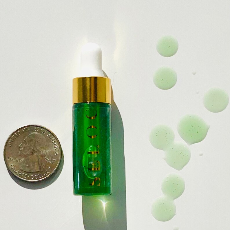 K&ouml;nnte beinhalten: Eine kleine gr&uuml;ne Glasflasche mit goldfarbenem Verschluss und einer Tropfpipette. Die Flasche ist mit "SOOCOO" beschriftet. Auf der wei&szlig;en Oberfl&auml;che befinden sich mehrere gr&uuml;ne Fl&uuml;ssigkeitstropfen.