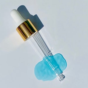 Pode incluir: Um frasco de vidro com conta-gotas, com uma tampa dourada e um l&iacute;quido azul pingando da ponta.