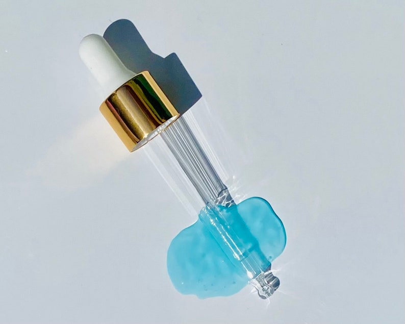 Pode incluir: Um frasco de vidro com conta-gotas, com uma tampa dourada e um l&iacute;quido azul pingando da ponta.