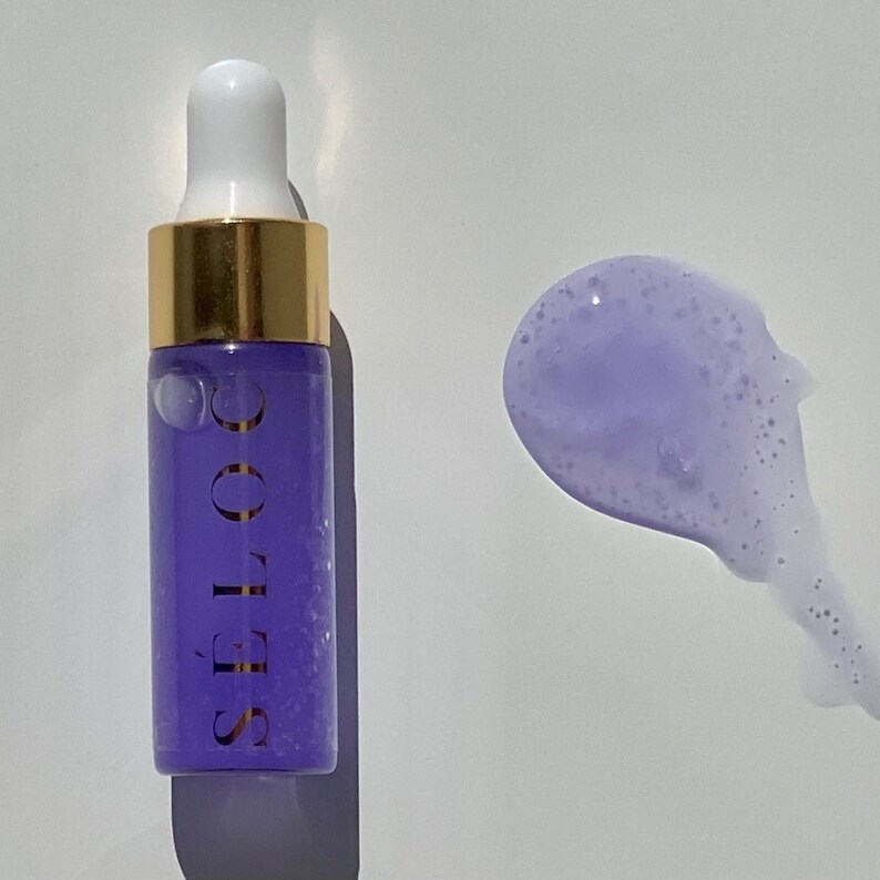 Pu&ograve; includere: Un piccolo flacone di vetro trasparente con un tappo dorato e un liquido viola all'interno. Il flacone &egrave; etichettato "S&Eacute;LO  CO.". Un liquido viola &egrave; stato versato su una superficie bianca.