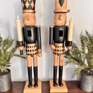 May include: Two wooden nutcracker candle holders. Each figure has a natural wood head and a black body with gold accents. One wears a crown, the other a hat. Both hold a cream-colored candle. The legs are black, and the base is natural wood.
