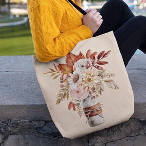 May include: A beige tote bag with a floral design featuring a bouquet of white, pink, and orange flowers in a brown vase. The flowers are surrounded by fall leaves and berries.