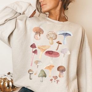 May include: A beige sweatshirt featuring a watercolour print of assorted mushrooms in shades of brown, red, yellow, and blue.