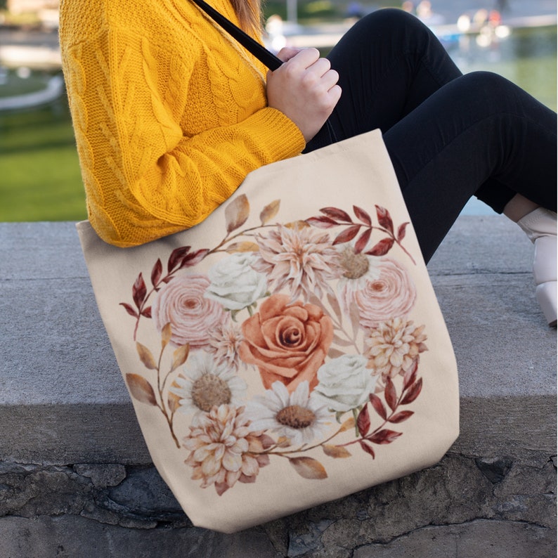 May include: A beige canvas tote bag featuring a floral design with a variety of flowers in shades of pink, white, and orange. The flowers are arranged in a circular pattern and surrounded by brown leaves.