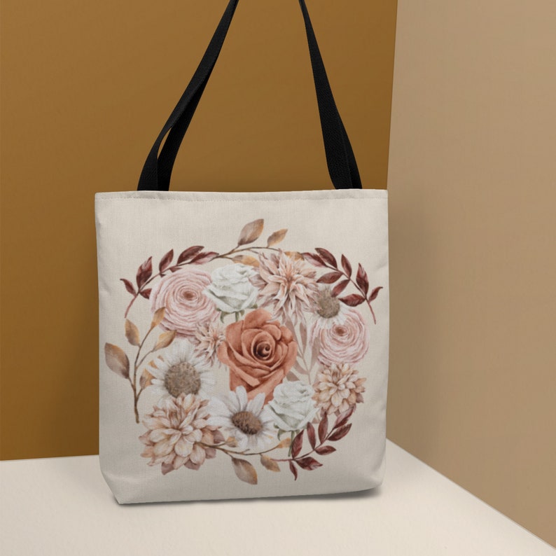 May include: A white tote bag with black straps featuring a floral design. The design includes pink, white, and orange roses, along with brown leaves and stems.