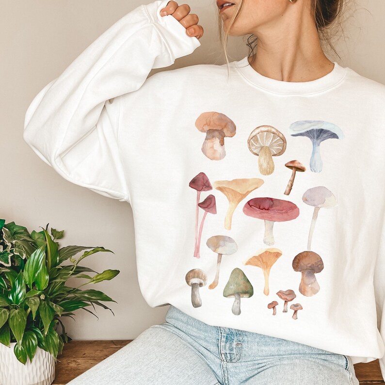 May include: White sweatshirt featuring a watercolour mushroom print. The mushrooms are in various colours, including red, orange, yellow, brown, and blue.