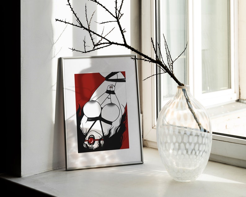 May include: A framed black and white illustration of a woman tied up with rope, wearing a red ball gag. The background is red. The frame is silver.