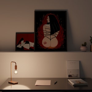 以下が含まれることがあります： 黒と赤の女性を描いた額装されたイラストが2つ。1つは背中に五芒星、もう1つは横たわっている。イラストは、ランプ、ノート、ペン、雑誌2冊が置かれたデスクの上の白い壁にある。