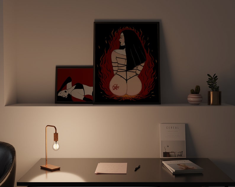 以下が含まれることがあります： 黒と赤の女性を描いた額装されたイラストが2つ。1つは背中に五芒星、もう1つは横たわっている。イラストは、ランプ、ノート、ペン、雑誌2冊が置かれたデスクの上の白い壁にある。
