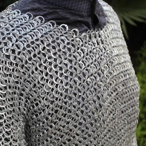 Peut inclure: Une chemise en cotte de mailles argentée composée de nombreux anneaux métalliques entrelacés. Le vêtement présente un motif circulaire dense et répétitif sur tout le torse et les épaules, porté sur une couche de base gris foncé texturée.