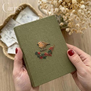 以下が含まれることがあります： 赤い実のついた枝にとまる鳥の刺繍が施された緑色のジャーナル。ジャーナルは人の手で持たれています。カバーは質感のある外観をしています。