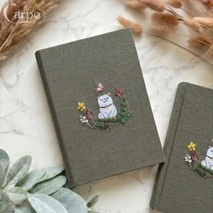 Puede incluir: Dos cuadernos verde oliva con diseños bordados. Uno presenta un gato blanco, una mariposa rosa y flores coloridas. El otro cuaderno tiene un diseño similar. El texto "carpe diem design" está en la esquina superior izquierda.