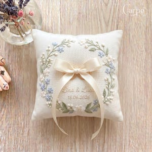 Peut inclure: Un coussin d'alliances carré blanc cassé orné d'une couronne florale brodée dans des tons de bleu, vert et blanc. Le centre affiche le texte "Lina & Luca" et "18.06.2026" avec un ruban de satin couleur crème noué pour maintenir deux anneaux.