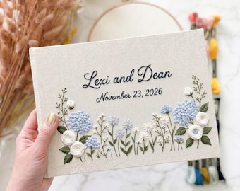 Può includere: Un album rettangolare in tessuto color crema con testo ricamato in nero personalizzato che recita Lexi and Dean e November 23, 2026. La copertina è decorata con grappoli di ortensie blu ricamati, fiori bianchi e foglie verdi.