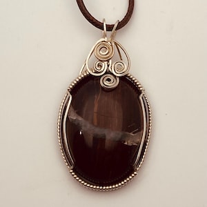 Può includere: Un ciondolo ovale in pietra preziosa marrone scuro con una venatura bianca orizzontale, racchiuso in una cornice di filo metallico color argento con spirali decorative sulla parte superiore, appeso a un semplice cordino marrone.