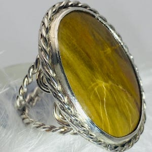 May include: A silver ring with a large, oval tiger's eye stone. The stone is a warm yellow-brown color with natural veining. The ring has a twisted rope design around the stone and on the band, creating a vintage or bohemian aesthetic.