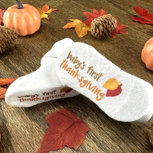May include: A pair of white baby socks with the text "Baby's first Thanksgiving" on them. The socks are decorated with a small orange and red leaf design.