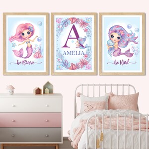 May include: Three framed prints with a blue background and a mermaid theme. The left print features a pink mermaid with the text "be brave". The middle print features a purple letter "A" with the name "Amelia" below it. The right print features a purple mermaid with the text "be kind".