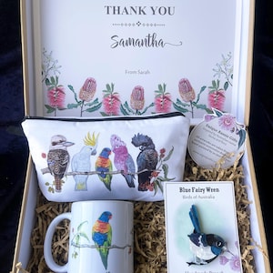 May include: A gift box filled with Australian themed gifts. The box contains a mug with a colourful bird illustration, a small pouch with a bird illustration, and a handmade brooch of a blue fairy wren. The box is decorated with shredded paper and a thank you card with the name Samantha.