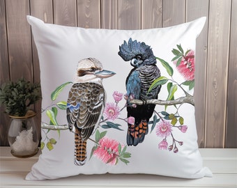 May include: White cushion featuring a black cockatoo and a kookaburra perched on a branch with pink flowers. The birds are painted in a realistic style.