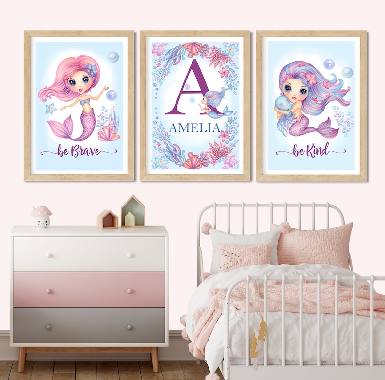 May include: Three framed prints with a blue background and a mermaid theme. The left print features a pink mermaid with the text "be brave". The middle print features a purple letter "A" with the name "Amelia" below it. The right print features a purple mermaid with the text "be kind".