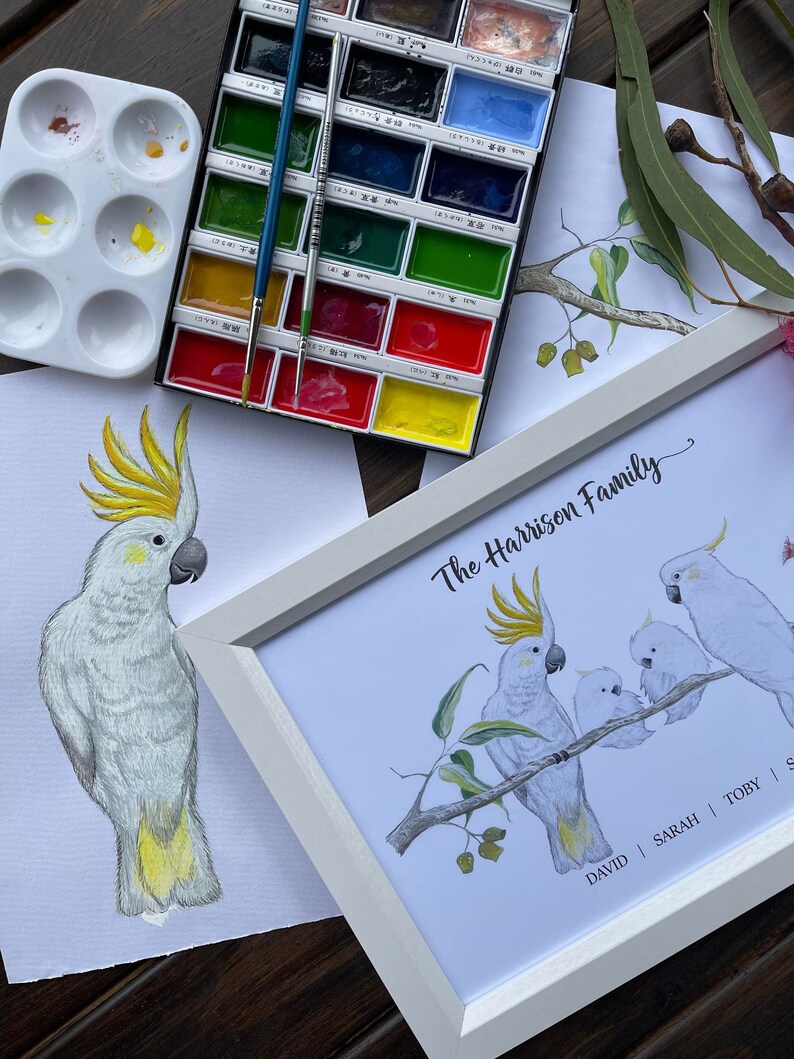 Pu&ograve; includere: Materiali per pittura ad acquerello e opere d'arte incorniciate con cacatua. La stampa incorniciata mostra una famiglia di cacatua appollaiati su un ramo, con il testo "The Harrison Family". &Egrave; visibile anche un dipinto ad acquerello di un singolo cacatua.