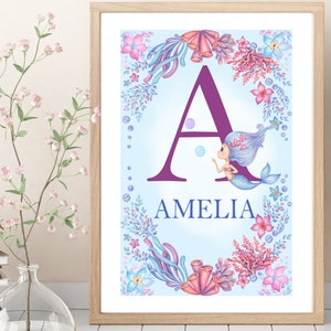 May include: A framed print with a purple letter A surrounded by a border of coral, seaweed, and flowers. A mermaid with blue hair is swimming next to the letter. The name Amelia is written below the letter.