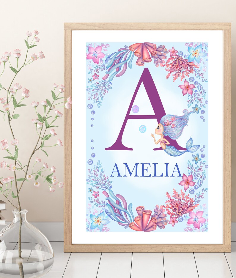 May include: A framed print with a purple letter A surrounded by a border of coral, seaweed, and flowers. A mermaid with blue hair is swimming next to the letter. The name Amelia is written below the letter.