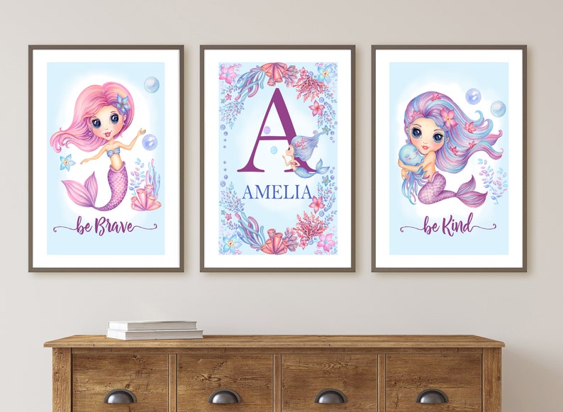 May include: Three framed prints with a mermaid theme. The left print features a pink mermaid with the text "be brave". The middle print features a purple letter "A" with the name "Amelia" and a small mermaid. The right print features a purple mermaid with the text "be kind".