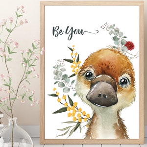 May include: A watercolor illustration of a platypus with a brown and white body, large black eyes, and a black beak. The platypus is surrounded by yellow and green flowers and leaves. The text "Be You" is written in black cursive above the platypus.