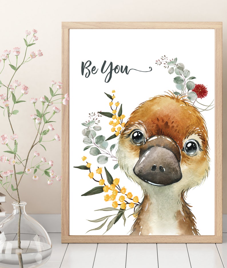 May include: A watercolor illustration of a platypus with a brown and white body, large black eyes, and a black beak. The platypus is surrounded by yellow and green flowers and leaves. The text "Be You" is written in black cursive above the platypus.
