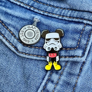 May include: A silver pin with the text "GAP DENIM EST. 1969" and a black and white enamel pin of a Mickey Mouse character wearing a Stormtrooper helmet. The pins are on a blue denim jacket.