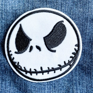 以下が含まれることがあります： 白い刺繍のパッチに、黒い糸で縫われた顔のデザインが施されています。口と目は縫い付けられています。この顔は、人気のハロウィーンキャラクターを彷彿とさせます。