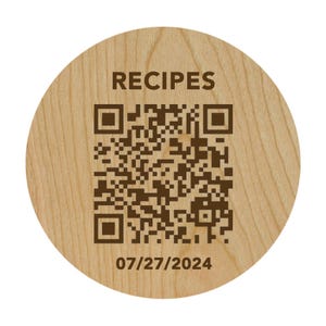 Op de afbeelding: Een rond houten voorwerp met het woord "RECIPES" bovenaan. Een QR-code staat in het midden, met de datum "07/27/2024" eronder. Het hout heeft een lichtbruine kleur en een zichtbare nerf.