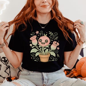 May include: Black t-shirt featuring a whimsical illustration of a smiling plant with pink flowers and a pot. The design includes decorative swirls, a skull, and small birds. The shirt is made of a soft material.