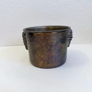 May include: A tarnished brass-toned metal pot with a cylindrical body and two decorative side handles. The surface shows a dark, aged patina with mottled brown and bronze tones throughout the exterior, featuring a smooth, wide circular rim.
