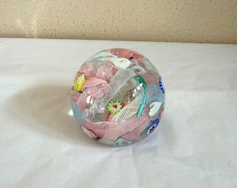 May include: A clear glass paperweight with colorful internal designs. The paperweight features pink, white, blue, and green decorative elements. The design includes floral and abstract patterns, creating a unique and artistic appearance.