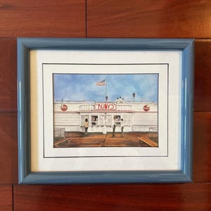 Könnte beinhalten: Ein gerahmtes Aquarellgemälde von Ruby's, einem Diner mit einer amerikanischen Flagge im Hintergrund. Das Diner ist weiß mit roten Akzenten und hat mehrere Personen davor stehen.