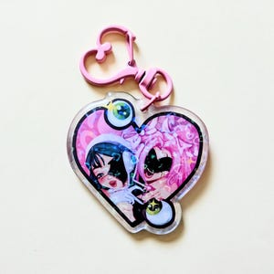 May include: A heart-shaped acrylic keychain featuring a pink and black illustration of two characters with stylized eyes. The keychain has a clear finish with a holographic effect and is attached to a pink plastic heart-shaped lobster clasp.