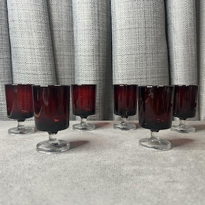 May include: Six ruby red wine glasses with clear stems and bases are arranged on a light gray surface. The glasses have a short, rounded bowl shape. The background features a textured, light gray curtain.