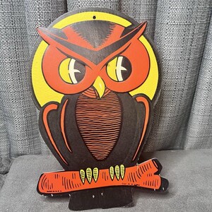 May include: A vintage Halloween owl decoration. The owl is black with orange and yellow accents, perched on an orange branch. The owl has large eyes and a beak. The decoration is made of cardboard and has a hole at the top for hanging.