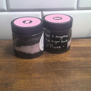 Puede incluir: Dos frascos de vidrio con tapas negras y etiquetas rosas. Los frascos contienen un exfoliante de azúcar marrón y rosa en capas. La etiqueta de un frasco dice "Type O Negative 4 oz Sugar Scrub Rose."