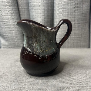 May include: A small, dark brown ceramic pitcher with a handle and spout. The pitcher has a glossy finish with a mottled green and brown glaze near the top. Gold script is visible on the side.
